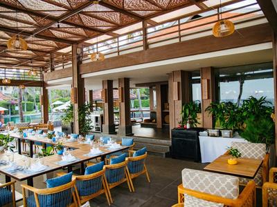 Offenes Restaurant mit Holzdach, blauen Stühlen, Holztischen und grünen Pflanzen im modernen Ambiente.