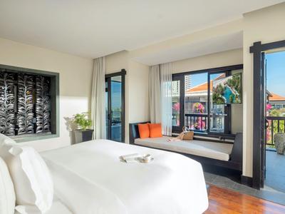 Jasny pokój hotelowy z dużym łóżkiem, siedziskiem przy oknie i balkonem z widokiem na miasto.