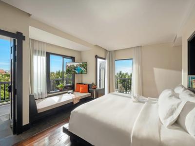 Camera d'hotel luminosa con letto grande, seduta alla finestra e vista su ambiente verde.