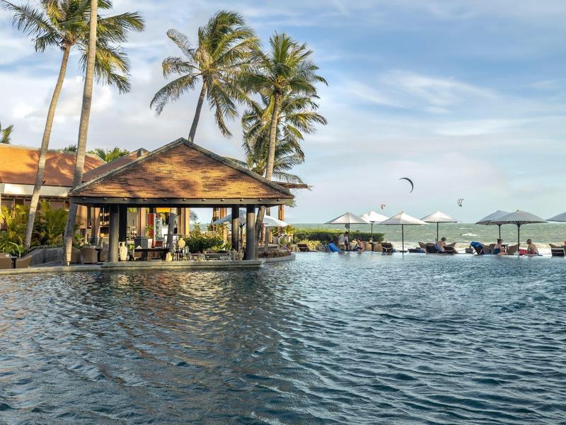 Duży basen z barem, palmami i parasolami przy morzu pod niebieskim niebem.