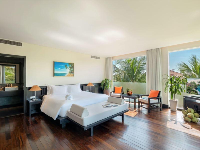 Jasny pokój hotelowy z podwójnym łóżkiem, miejscem do siedzenia i dużym oknem z widokiem na palmy.