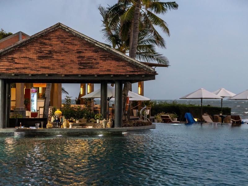 Wieczorne ujęcie stylowego baru przy basenie z oświetlonymi miejscami do siedzenia i widokiem na morze.