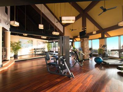 Světelná fitness místnost s dřevěnou podlahou, moderními stroji, velkými okny a rostlinami.