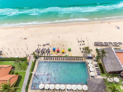 Vue aérienne d'une piscine de complexe avec parasols, chaises longues et vagues de l'océan en arrière-plan.