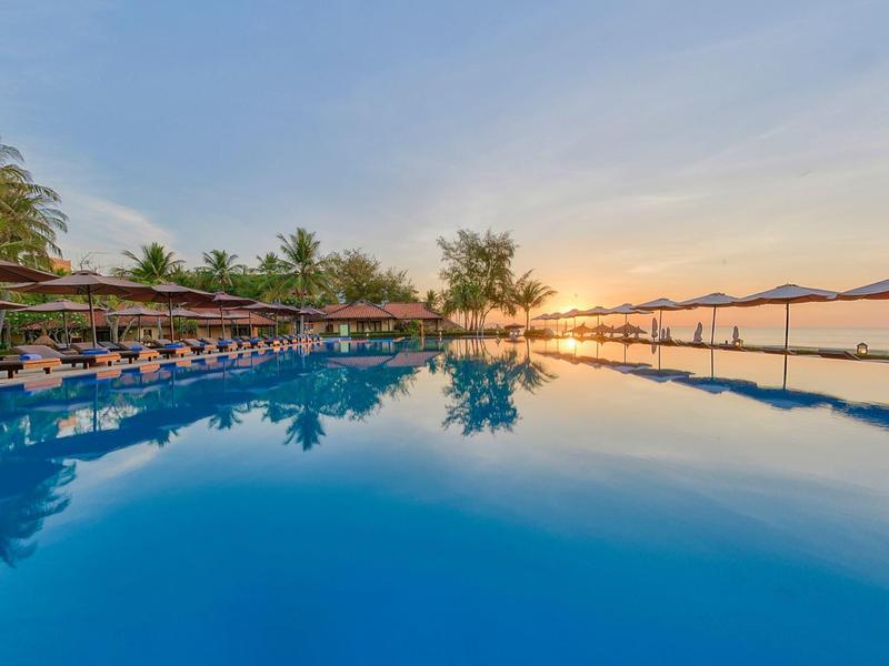 Une grande piscine au coucher du soleil avec des chaises longues et des palmiers à côté d'un hôtel.