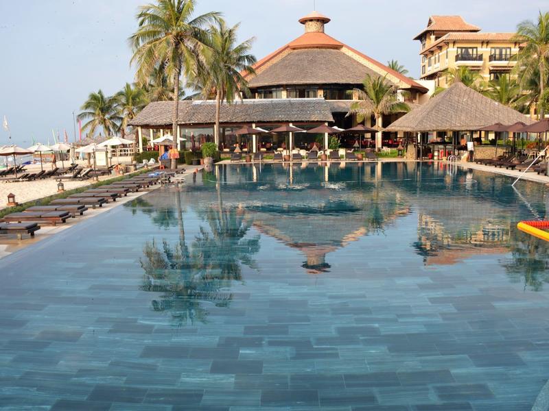 Grande piscine avec palmiers et bâtiments du complexe en arrière-plan sous un ciel clair.