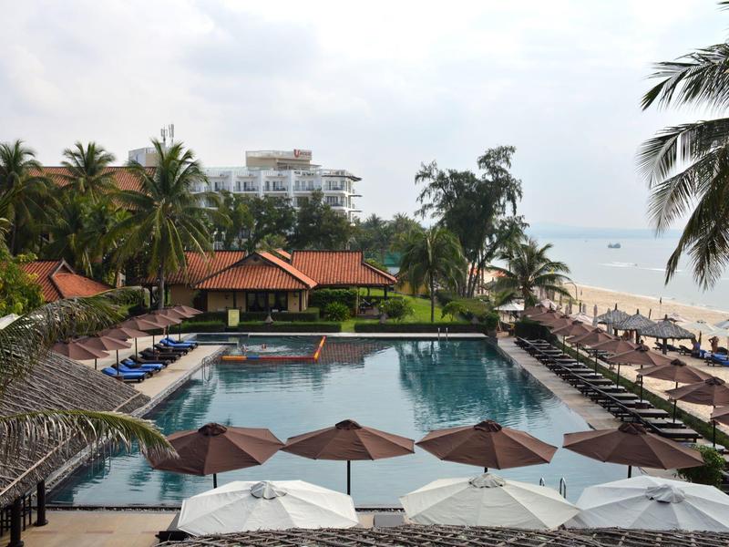 Piscine de la station avec parasols, palmiers et transats sur la plage sous un ciel nuageux.