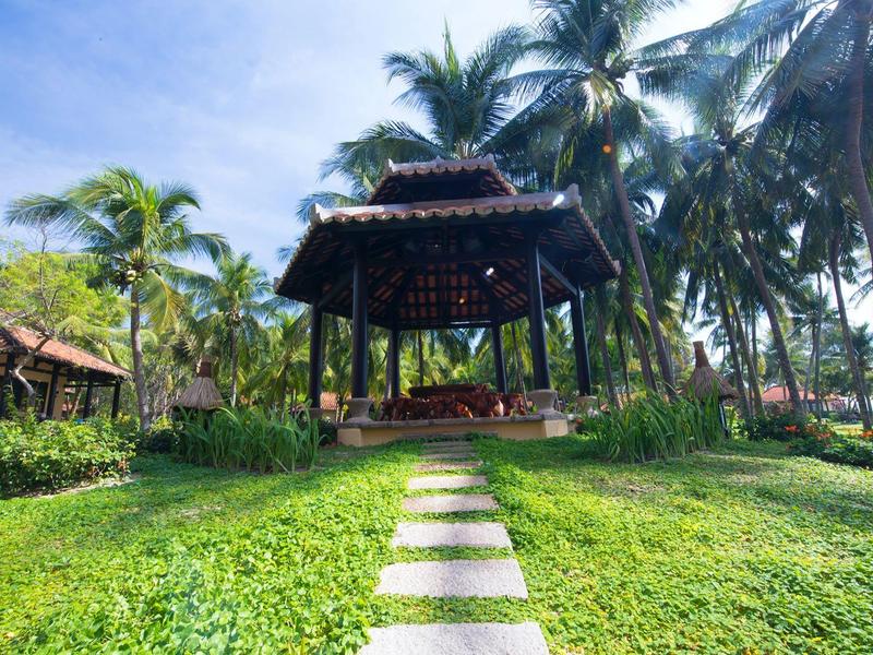 Pavillon avec sièges en bois entouré de palmiers et d'herbe verte sous un ciel bleu.