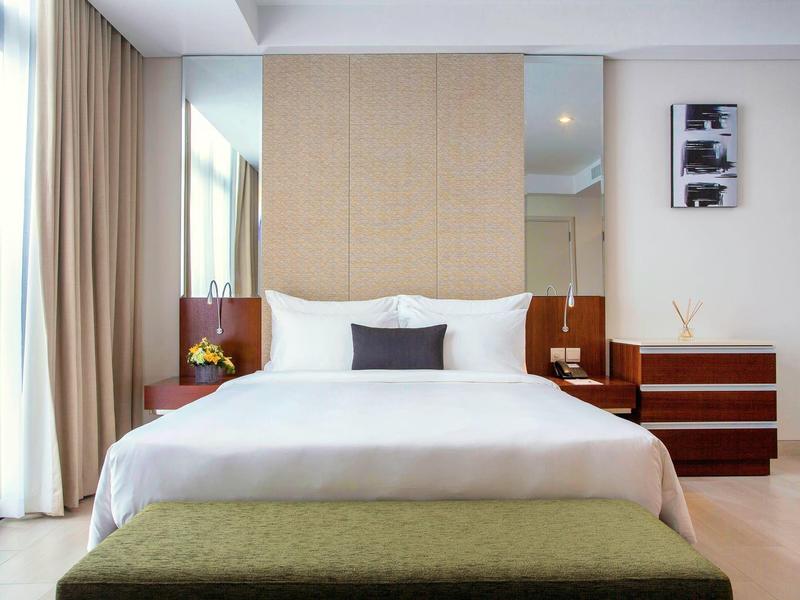 Modernes Schlafzimmer mit großem Bett, weißer Bettwäsche, zwei Nachttischen und grünem Hocker vorne.