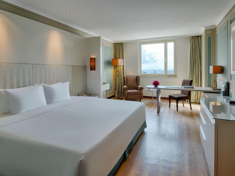 Helles Hotelzimmer mit großem Bett, Sessel, Schreibtisch und großen Fenstern, helles Holzfußboden.