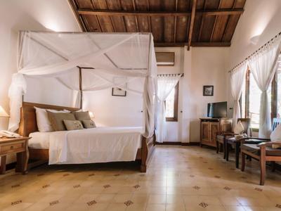 Chambre d'hôtel lumineuse avec lit à baldaquin, plafond en bois et coin salon confortable près de la fenêtre.