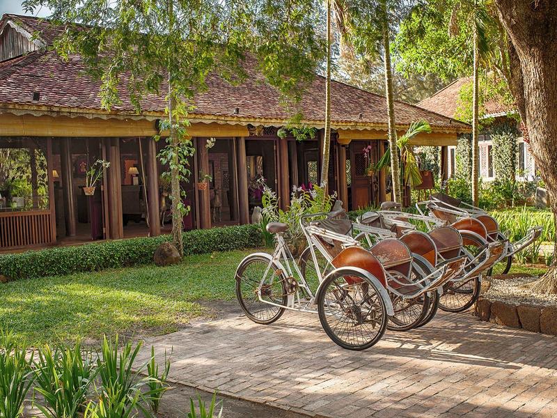 Vélos et pousse-pousse devant un bâtiment d'hôtel avec jardin et arbres.