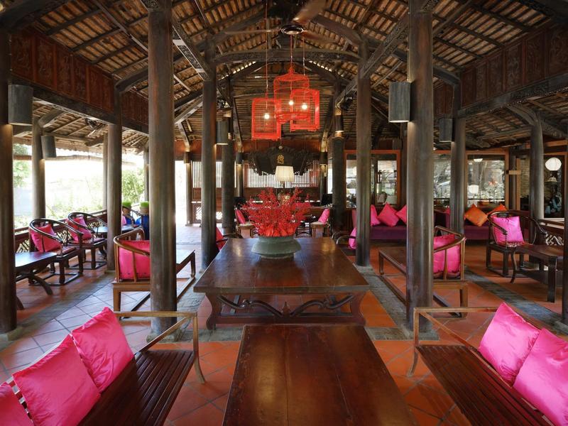 Salle à manger rustique ouverte avec tables en bois et coussins roses, entourée de poutres en bois.