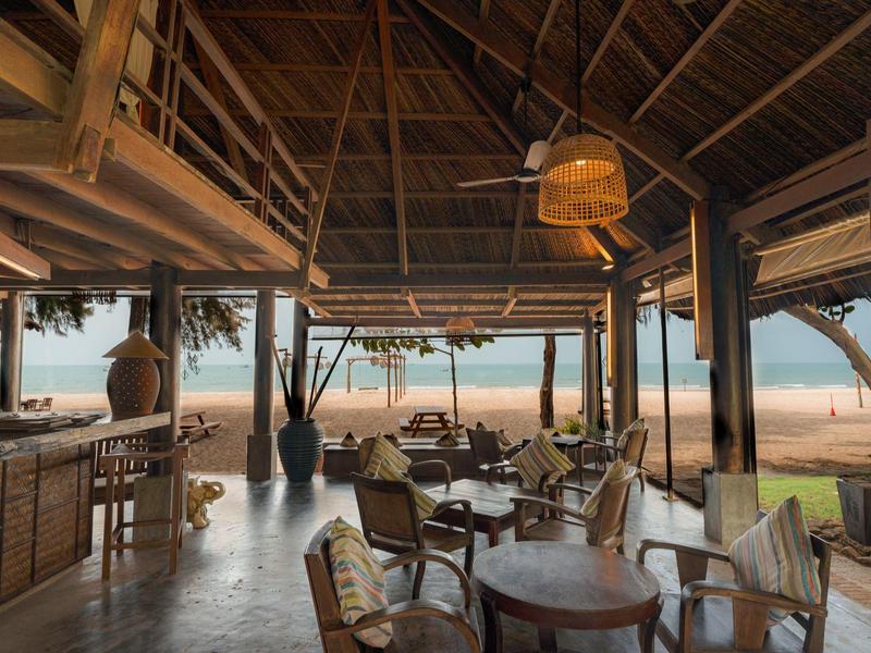 Bar de plage ouverte avec plafond en bois, tables et chaises donnant sur le sable et la mer.