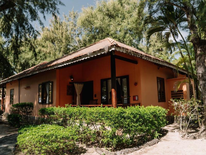 Une maison orange au toit en tuiles rouges, entourée de buissons et d'arbres par temps ensoleillé.