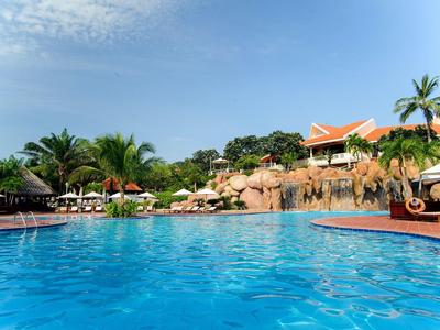 Gran piscina de hotel con sombrillas, palmeras y edificios bajo un cielo azul.