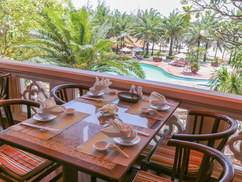 Mesa en balcón con desayuno dispuesto y vista a piscina tropical y palmeras en hotel.