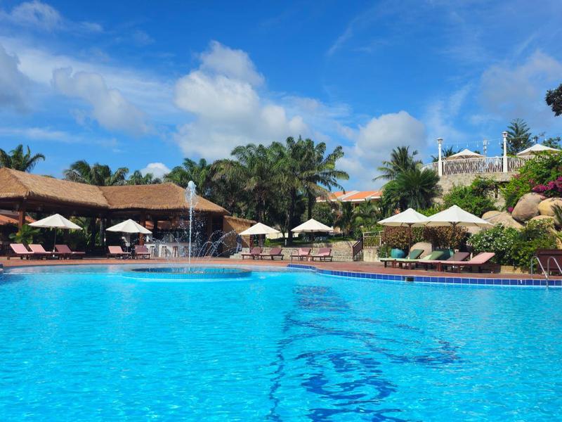 Gran piscina exterior con agua azul, rodeada de sombrillas y palmeras en un día soleado.