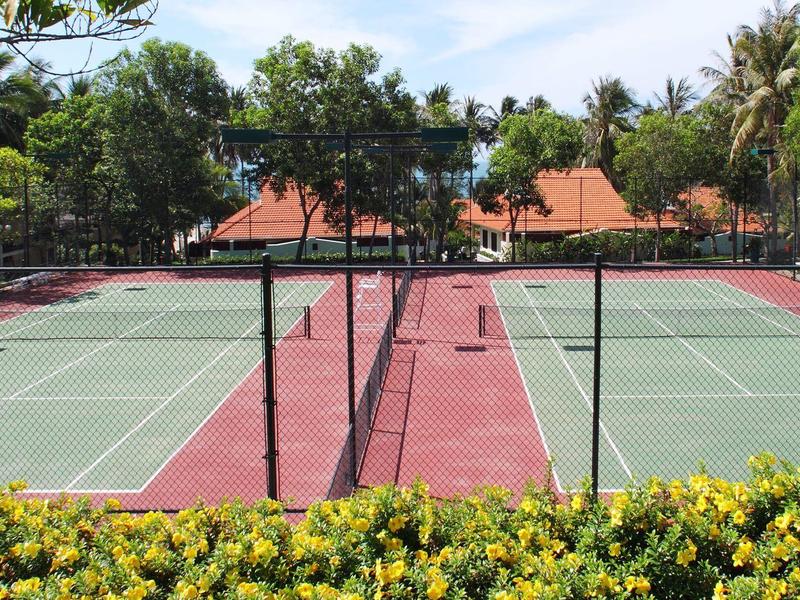 Dos canchas de tenis con suelo rojo y verde, rodeadas de árboles y flores amarillas al frente.