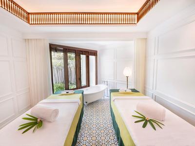 Heller Wellnessraum mit zwei Liegen, grünen Akzenten, großer Fensterfront und Badewanne im Hintergrund.