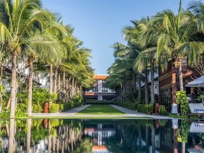 Ein langer Pool mit Palmen beidseits, der zu einem Hotelgebäude unter blauem Himmel führt.