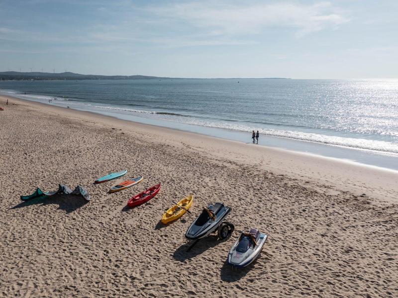 Strand mit sieben unterschiedlichen Kajaks in Reihen, zwei Personen am Wasser, ruhiges Meer und Himmel.