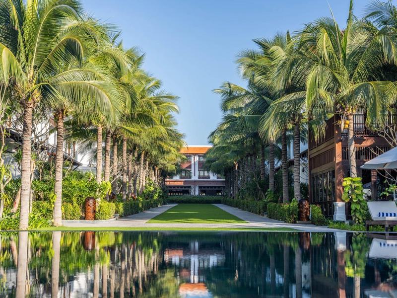 Ein langer Pool mit Palmen beidseits, der zu einem Hotelgebäude unter blauem Himmel führt.
