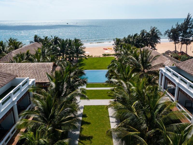 Blick auf tropische Gartenanlage mit Pool zwischen Gebäuden, Palmengärten und Strand im Hintergrund.