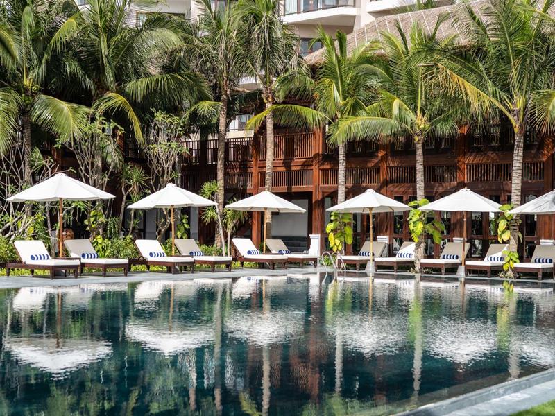 Großer Pool mit weißen Sonnenschirmen, Liegestühlen und Palmen vor einem Hotelgebäude.