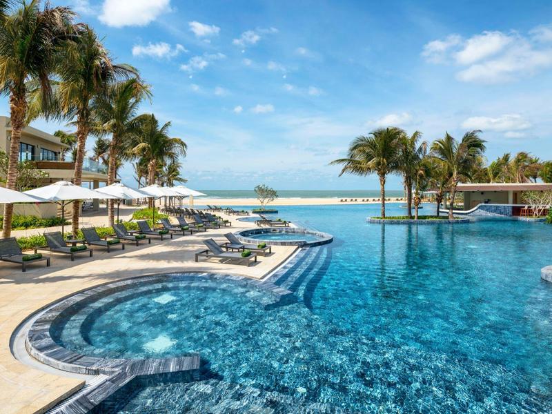 Luxusní bazén s lehátky a palmami pod světle modrou oblohou.