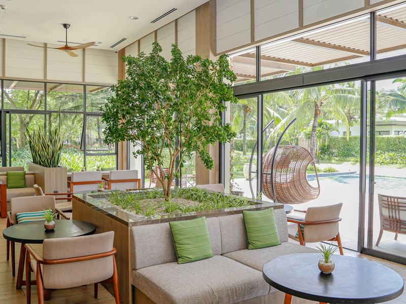 Světlý salón s velkými okny, béžovou pohovkou a zelenými rostlinami v moderním hotelu.