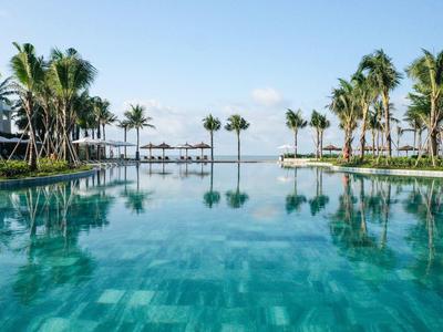 Una grande piscina con acqua limpida circondata da palme e lettini, con vista sul mare.