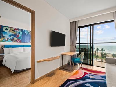 Lichte hotelkamer met tweepersoonsbed, tv, bureau en balkon met zeezicht.