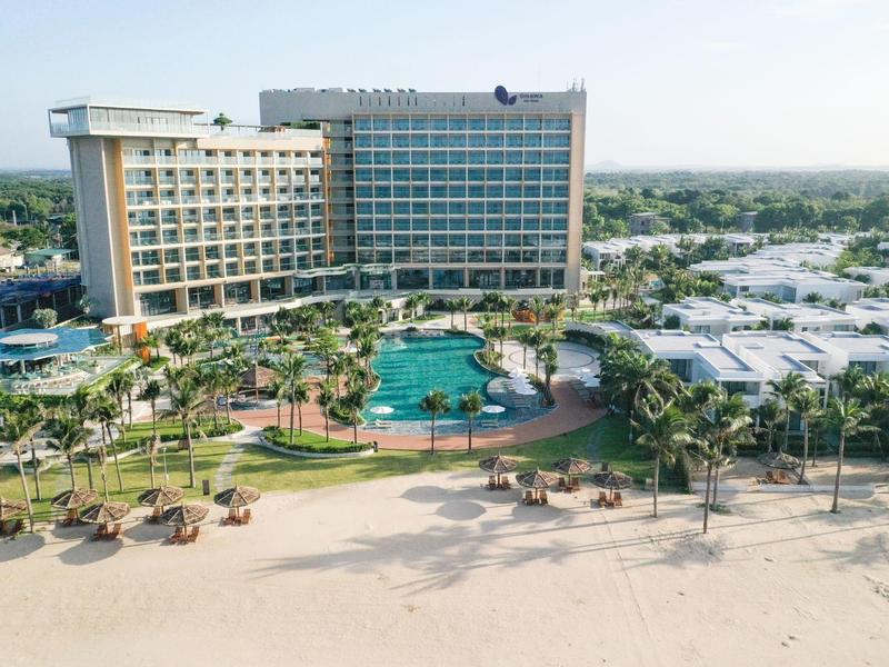 Luxe hotel met groot zwembad en palmbomen direct aan het zandstrand onder heldere lucht.
