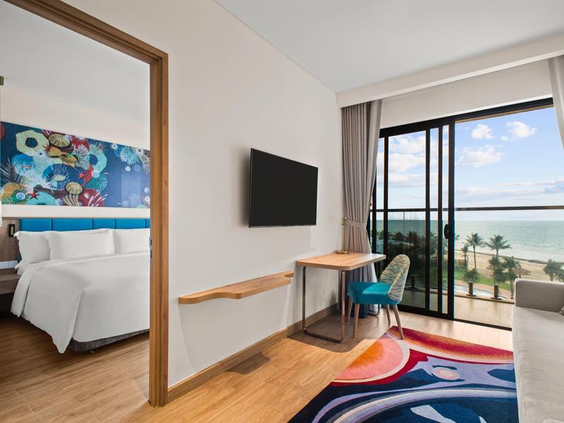 Lichte hotelkamer met tweepersoonsbed, tv, bureau en balkon met zeezicht.