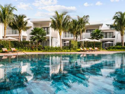 Complesso alberghiero moderno con piscina, circondato da palme e piante fiorite sotto un cielo azzurro.