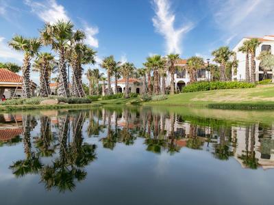 Hotel con palme e lago che riflette sotto un cielo blu.
