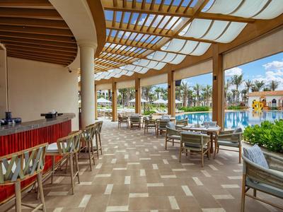 Lounge esterna coperta con mobili in legno accanto a piscina e edifici dell'hotel.