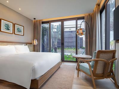 Modern hotel room with large bed, chair, and garden view through glass windows.