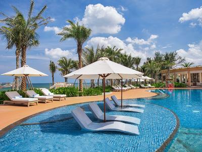Pool area with lounge chairs in water, sun umbrellas, and palm trees at a resort in sunny weather.