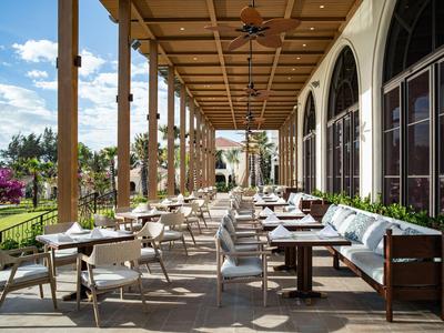 Covered hotel terrace with tables, chairs, and flower pots under a blue sky.