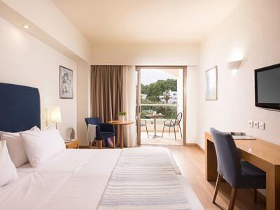 Habitación de hotel luminosa con cama doble, balcón, mesa con sillas y televisor en la pared.