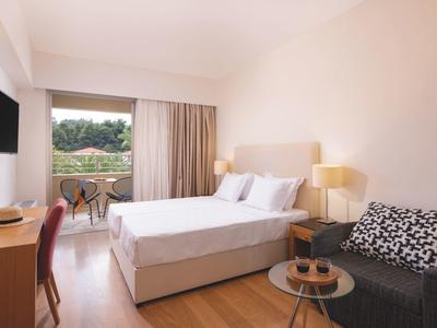 Habitación de hotel luminosa con cama doble, sofá, escritorio, balcón y vistas a un paisaje verde.