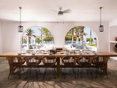 Gran mesa de comedor con sillas en un espacio abierto con arcos y vista a palmeras afuera.