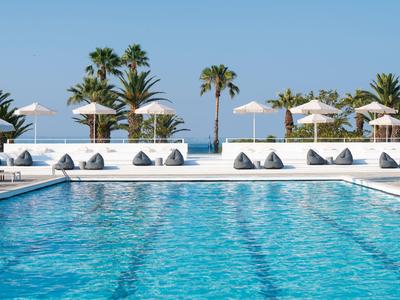 Piscine d'hôtel bleue avec parasols blancs, coussins gris, palmiers et vue sur la mer.
