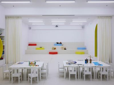 Espacio de juego infantil moderno y blanco con pequeñas mesas y cojines de colores en la pared.