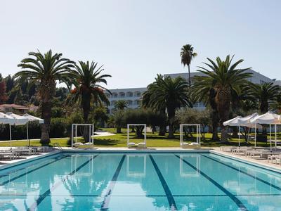 Piscine extérieure devant un hôtel, entourée de palmiers et de parasols au bord de l'eau.