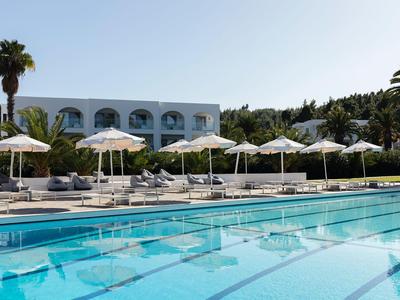 Piscine d'hôtel moderne avec chaises longues et parasols blancs devant un bâtiment blanc sous un ciel bleu clair.