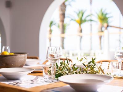 Table élégante avec verres et branches d'olivier devant des fenêtres éclairées par le soleil et des palmiers.