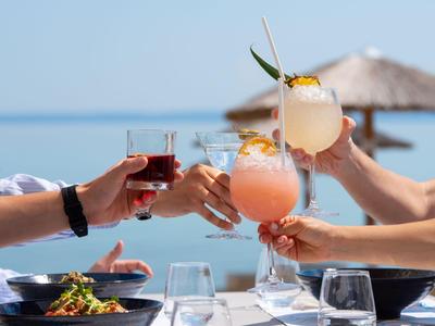 Plusieurs personnes trinquent avec différentes boissons devant une vue sur la mer et un parasol en paille.